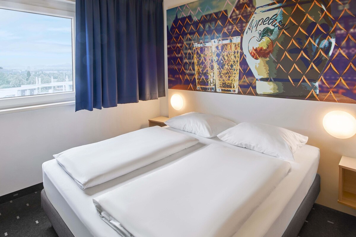 A hotel room with a bed, two bedside tables, a window with curtains, and a wall mural.