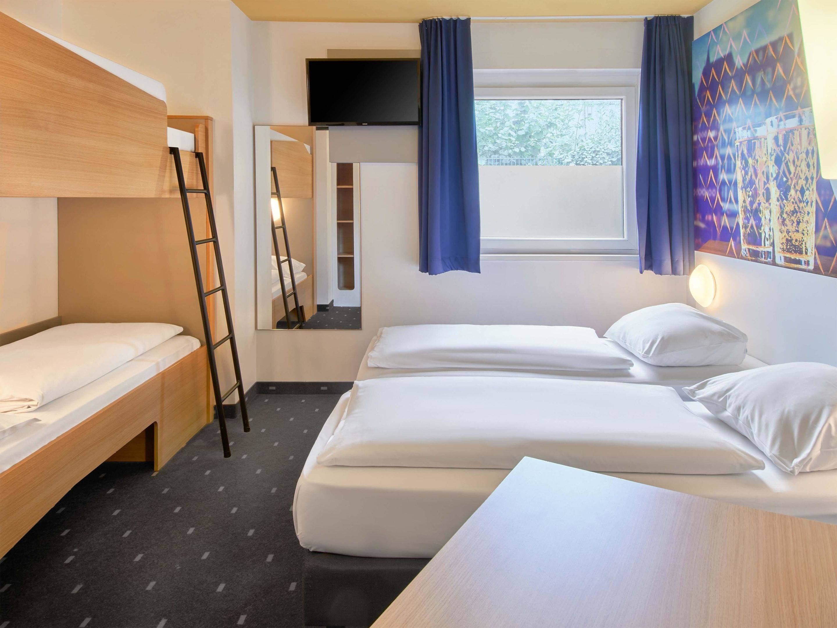 Een hotelkamer met twee bedden, een houten bureau, een ladder, een raam met gordijnen en een schilderij aan de muur.