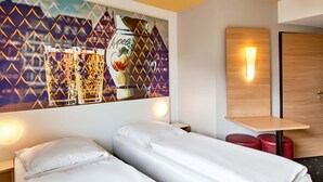 A hotel room with two single beds, a small table, a red stool, and a wall-mounted light.