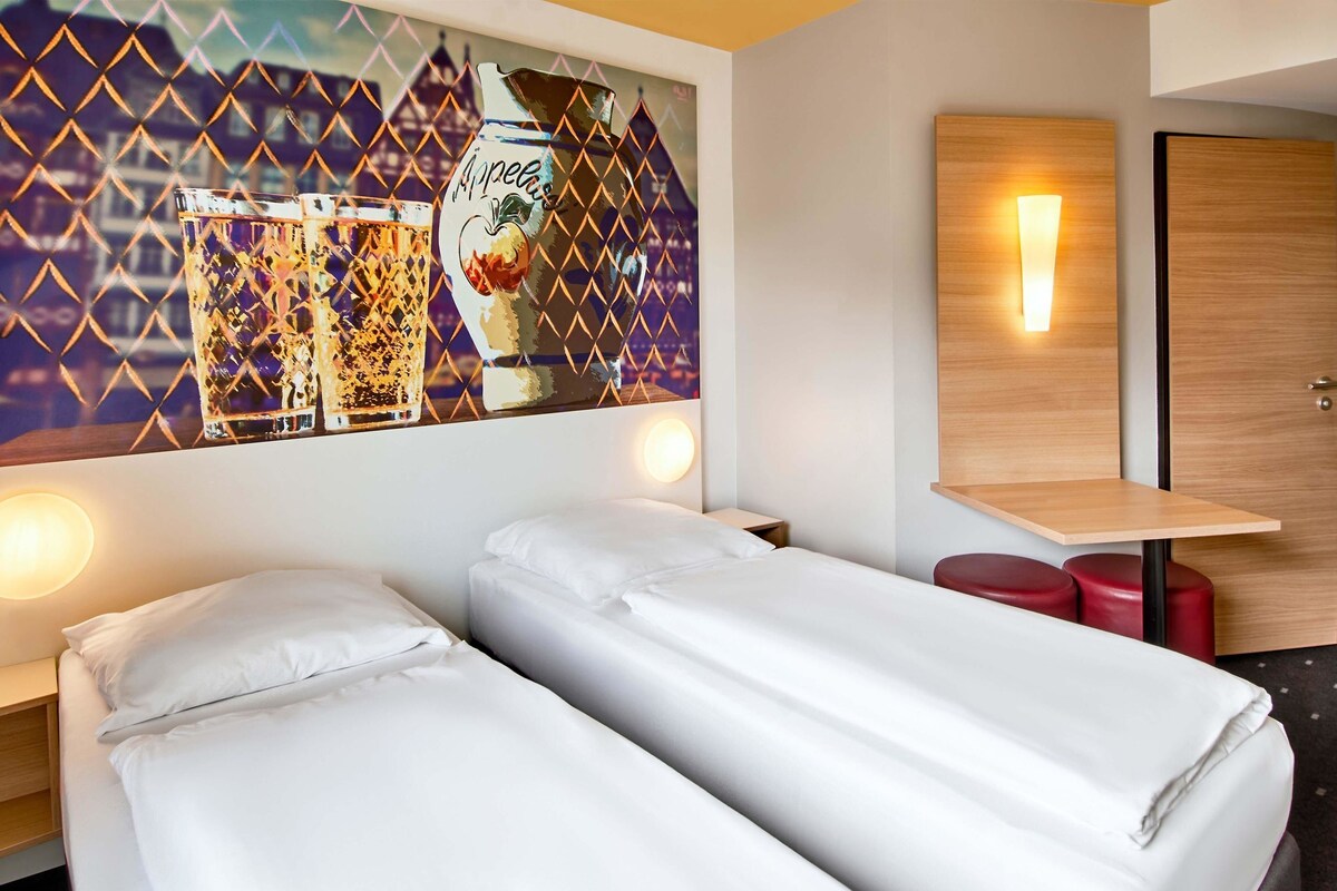 A hotel room with two single beds, a small table, a red stool, and a wall-mounted light.