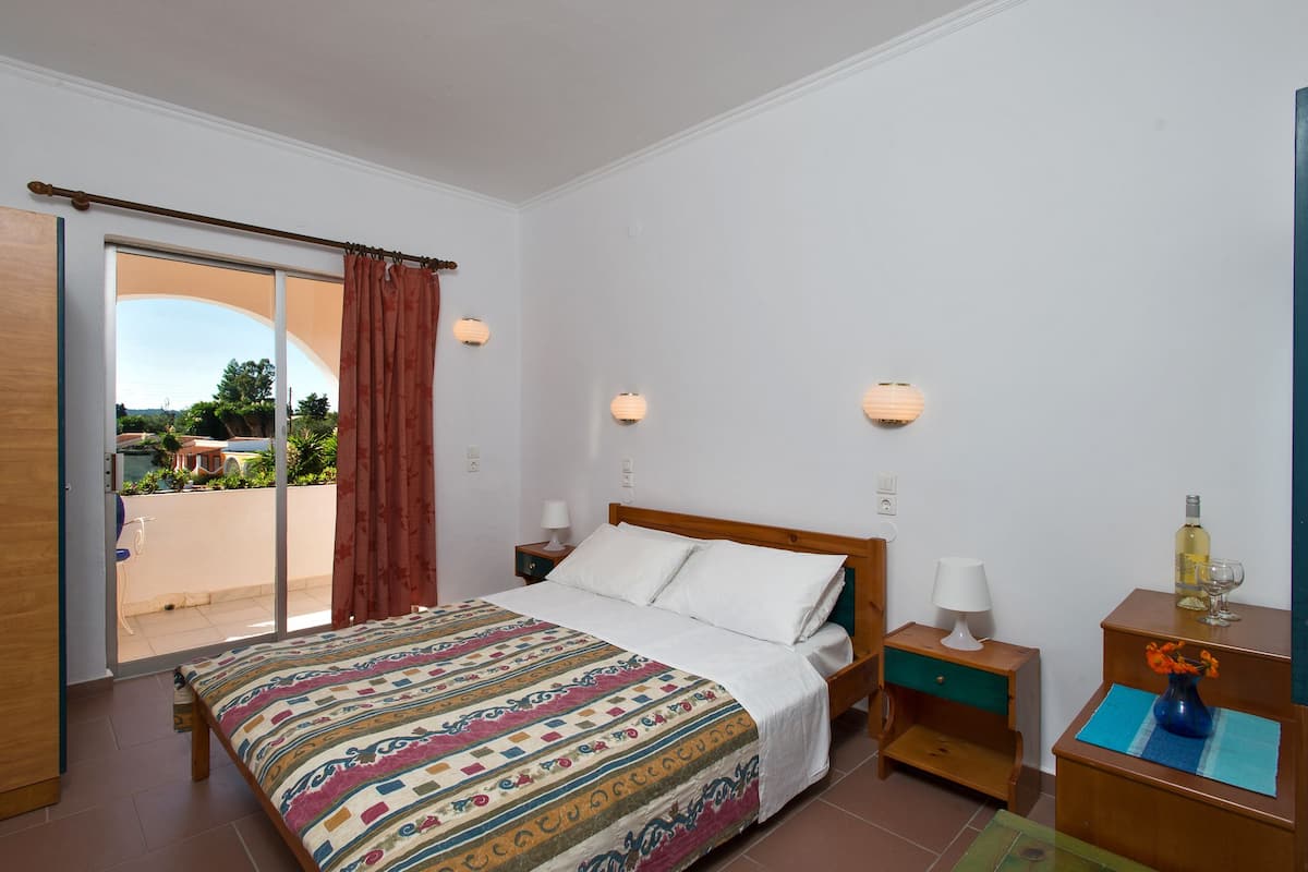 A bedroom with a bed, a nightstand, a lamp, a vase with flowers, and a view of the outdoors.