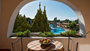 A balcony with a view of a swimming pool, a tall tree, and a table with a fruit basket.