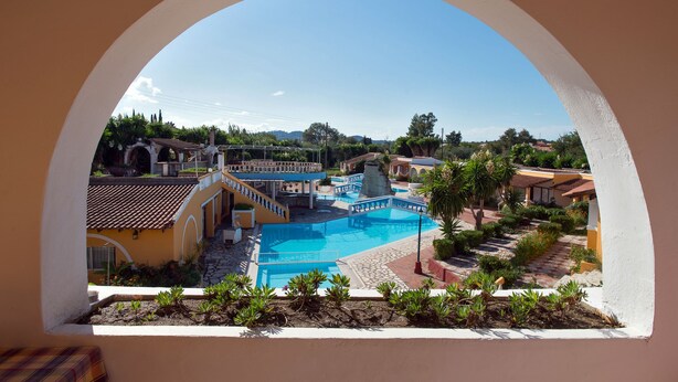 A view through an arched window of a pool area with a clear blue sky, surrounded by buildings with terracotta roofs and a well-maintained garden.