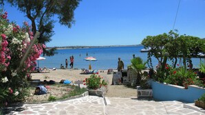 A beachside patio with stone flooring, surrounded by potted plants and flowers, overlooking a calm sea with people swimming and sunbathing. - Studios Diogenis (Naxos)