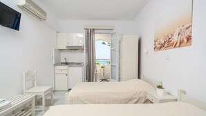 A hotel room with a bed, a desk, a chair, a TV, a kitchen area, and a view of the sea. - Studios Diogenis (Naxos)