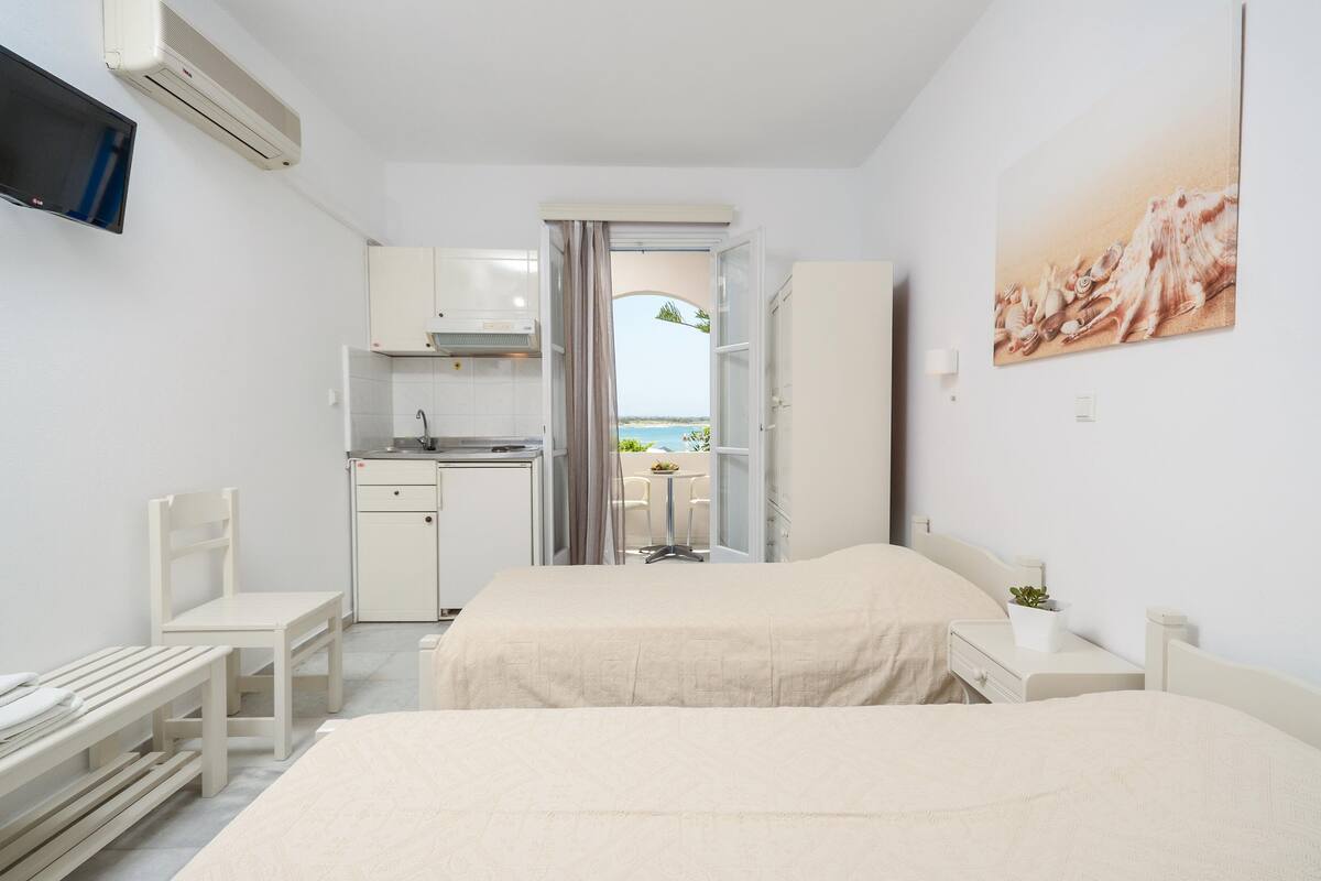 A hotel room with a bed, a desk, a chair, a TV, a kitchen area, and a view of the sea.