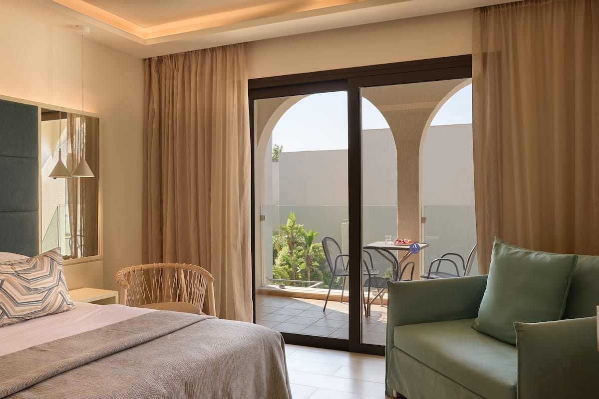A hotel room with a bed, a green armchair, a dining table, and a sliding glass door leading to an outdoor area with chairs and a table.