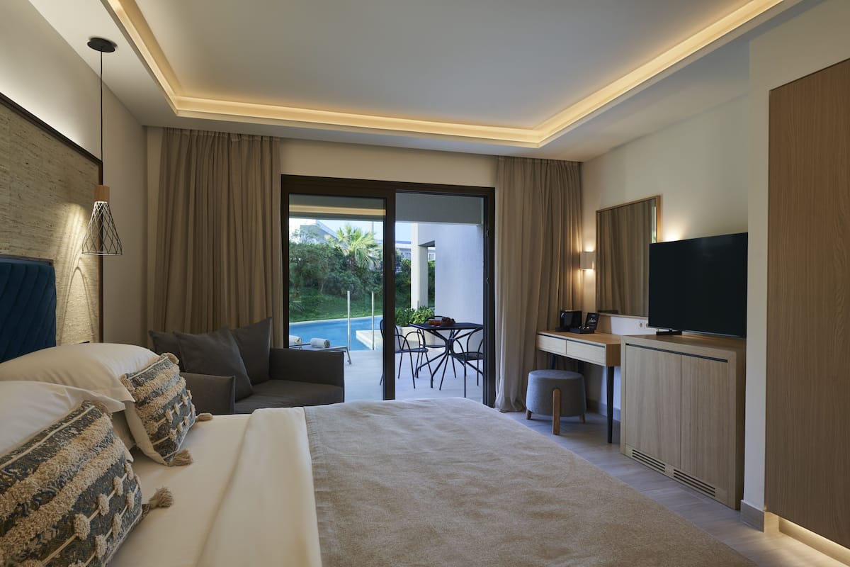 A modern hotel room with a bed, a desk, a television, and a view of a pool and greenery.