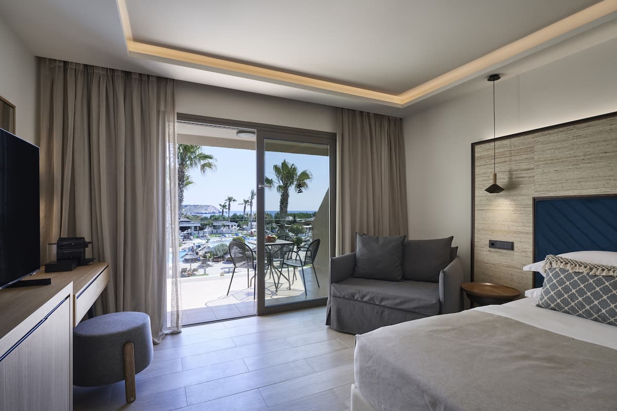 A modern hotel room with a balcony, a bed, a desk with a TV, and a seating area with a table and chairs.
