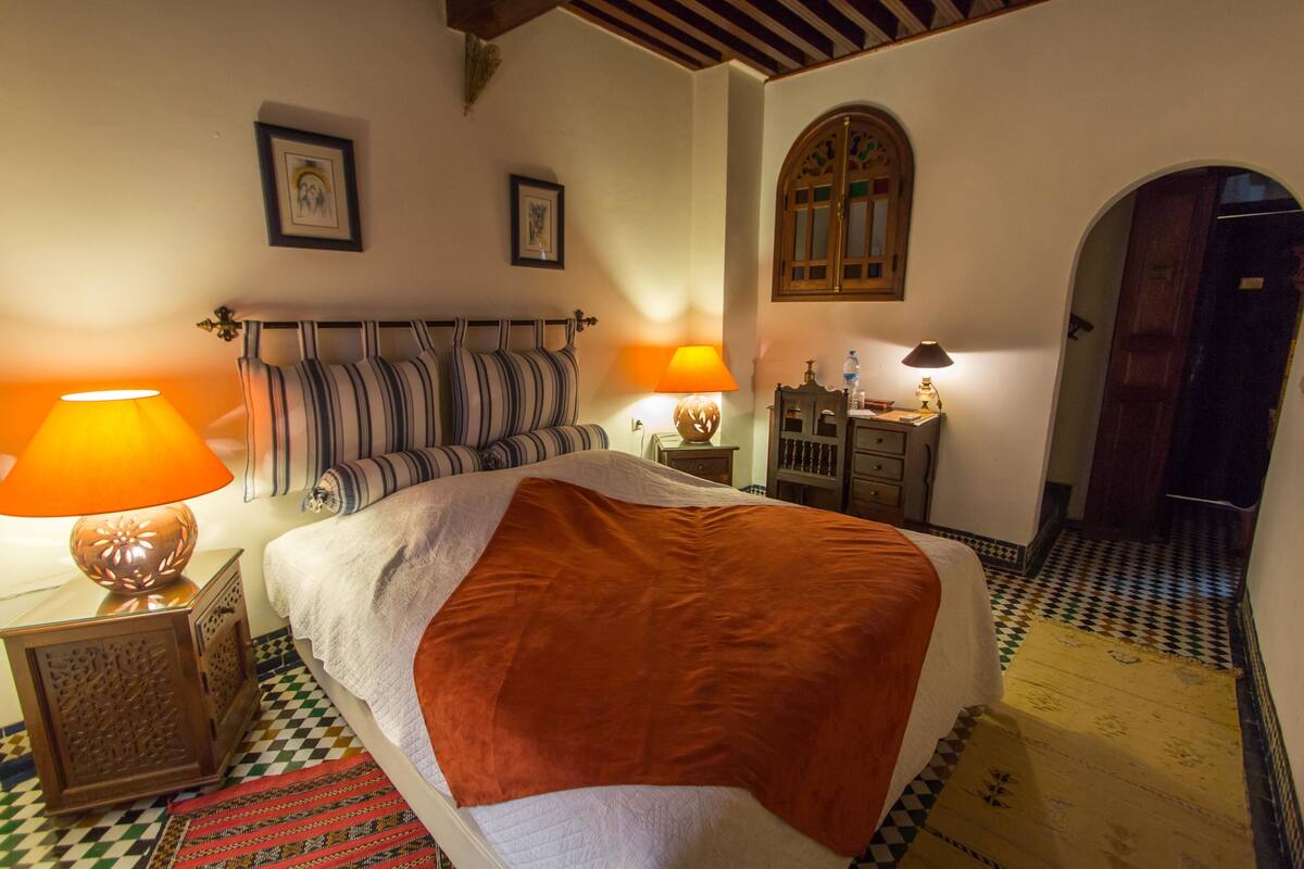 A bedroom with a bed, two bedside lamps, a striped headboard, a wooden cabinet, and a checkered floor.