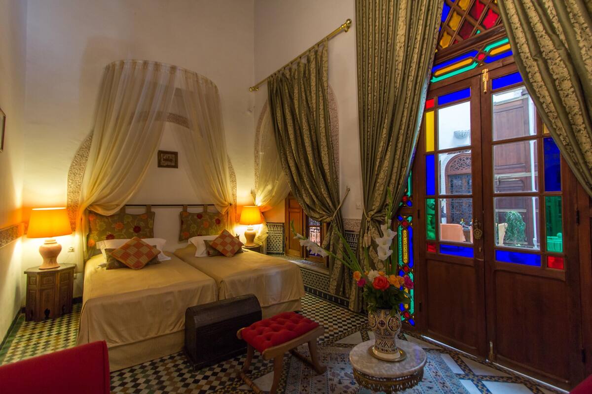A room with two beds, a red bench, a lamp, and a vase with flowers.