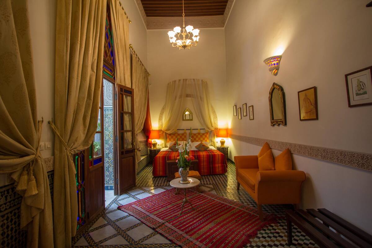 A room with a bed, a sofa, a chandelier, and a patterned rug.