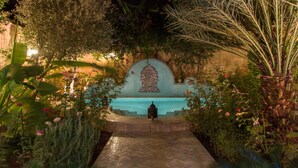 A nighttime view of a pool area with a tiled floor leading to a pool, surrounded by lush greenery and a decorative wall with a patterned design. - Riad Norma (Fes)