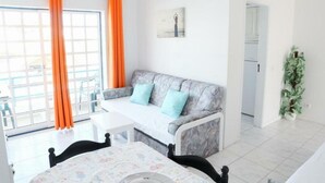 A room with a sofa, a dining table, and a window with orange curtains. - Sereia da Oura (Albufeira)
