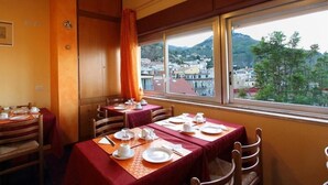A dining area with tables set for four, each with white plates, cups, and cutlery. The room has wooden walls and a large window offering a view of a town and hills. - Inn Piero (Taormina)