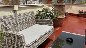 A wicker sofa and table on a rooftop terrace with potted plants and a view of a town and mountains. - Inn Piero (Taormina)