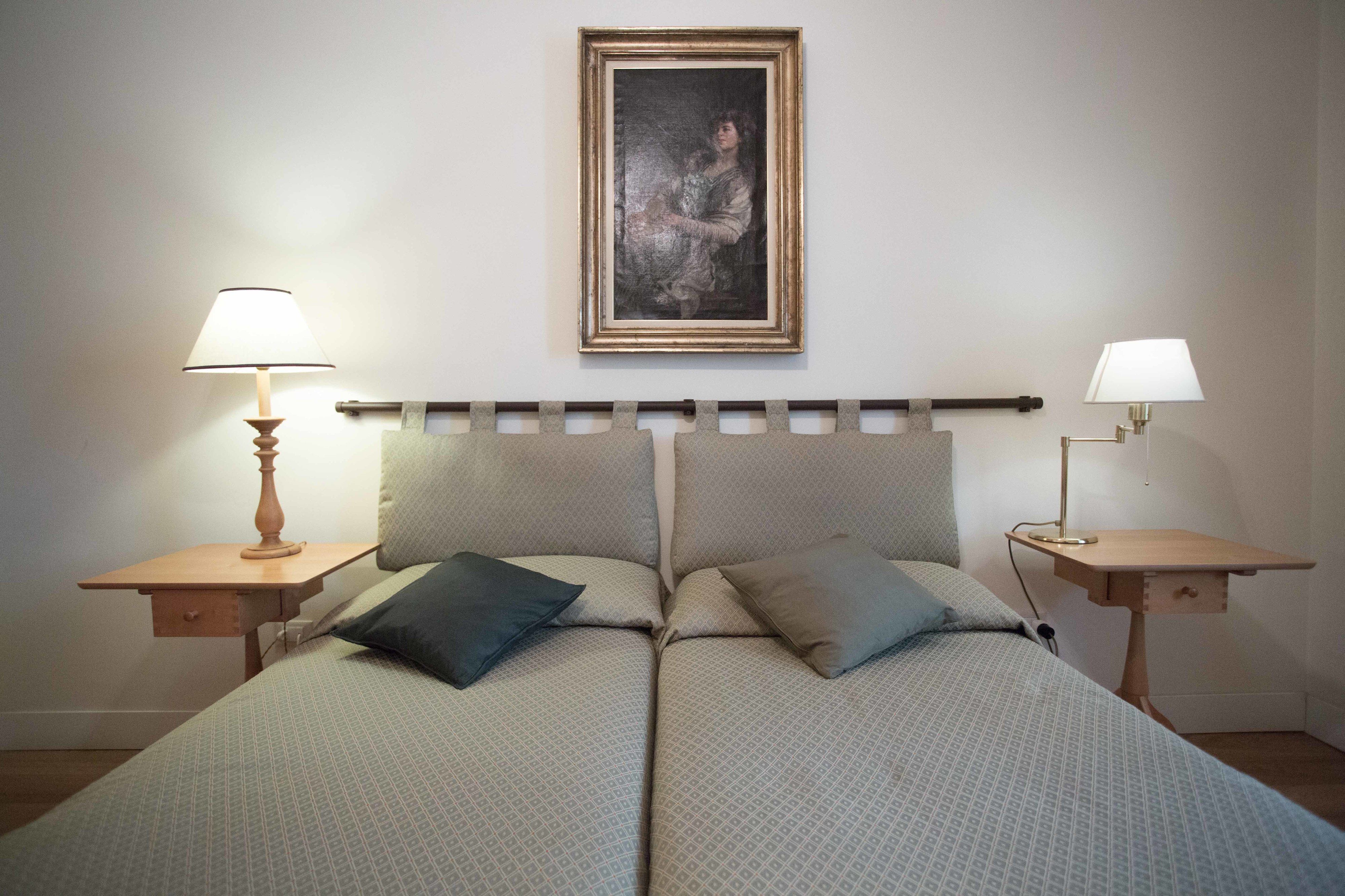 Uma cama de casal com cabeceira, duas mesas de cabeceira com abajures e um quadro na parede.