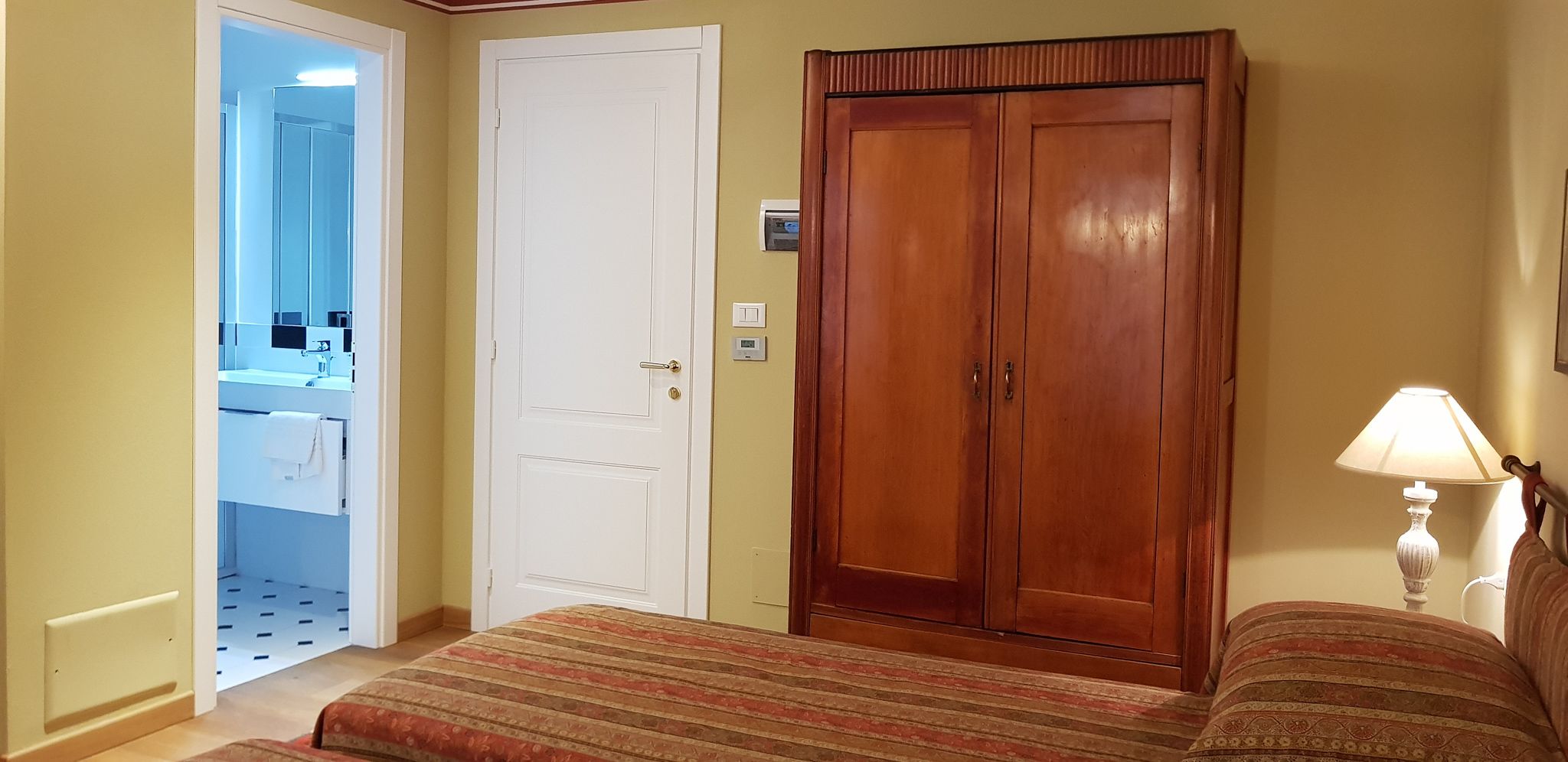 Un dormitorio con cama, armario, baño visible a través de una puerta abierta y lámpara de noche.