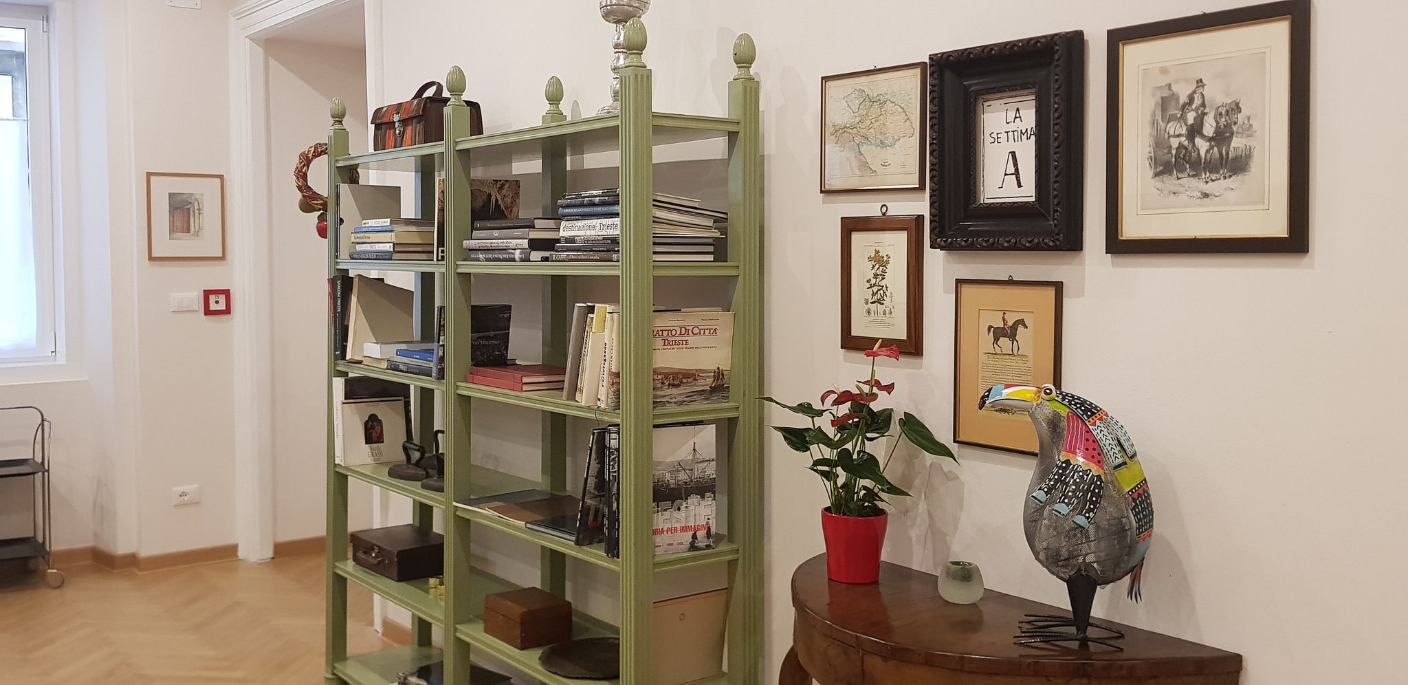 Une pièce avec une bibliothèque verte remplie de livres, un vase rouge contenant des fleurs et une sculpture d’oiseau colorée posée sur une table en bois.