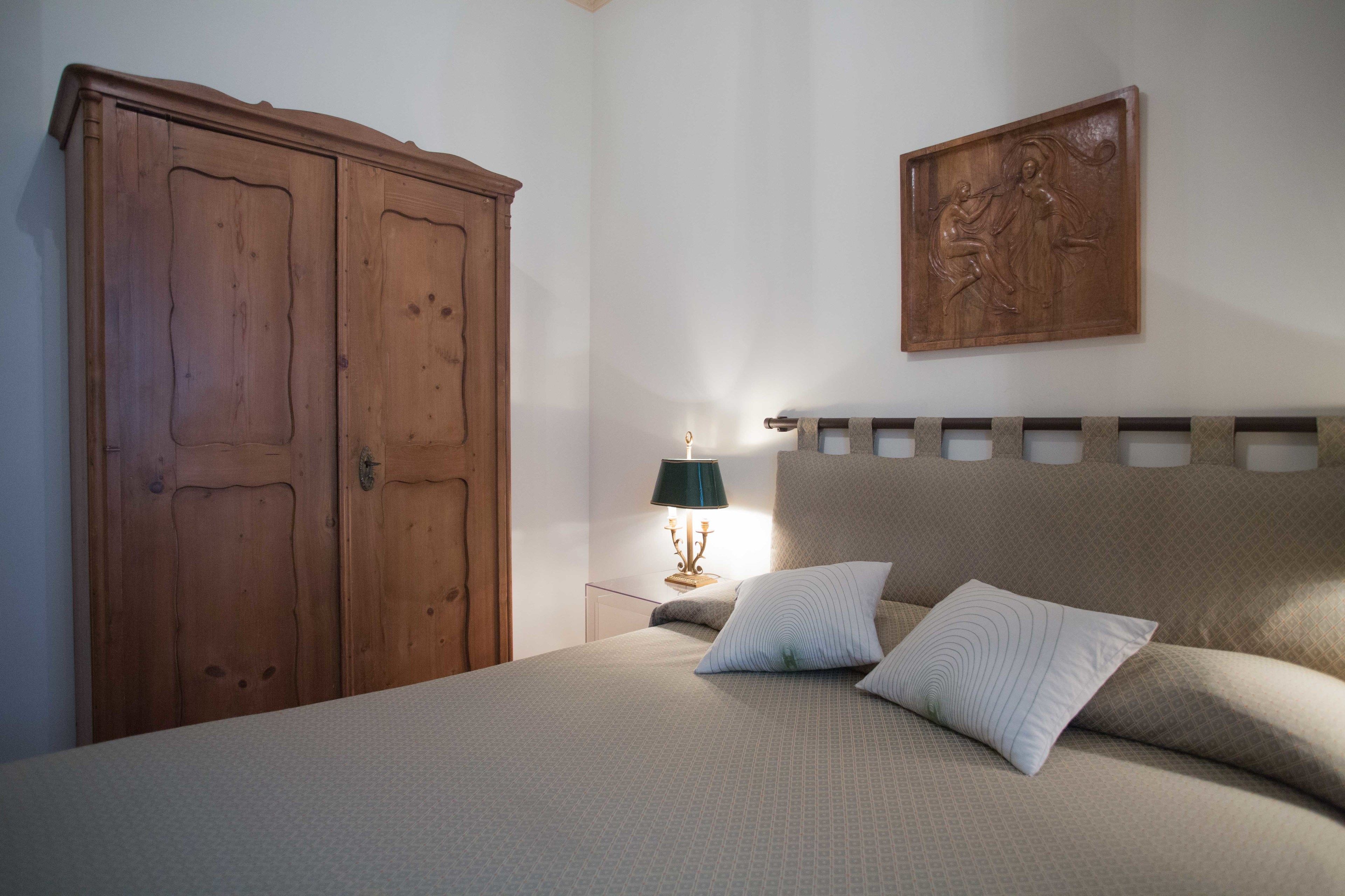 A bedroom with a bed, pillows, a wooden wardrobe, a nightstand with a lamp, and a wall-mounted artwork.
