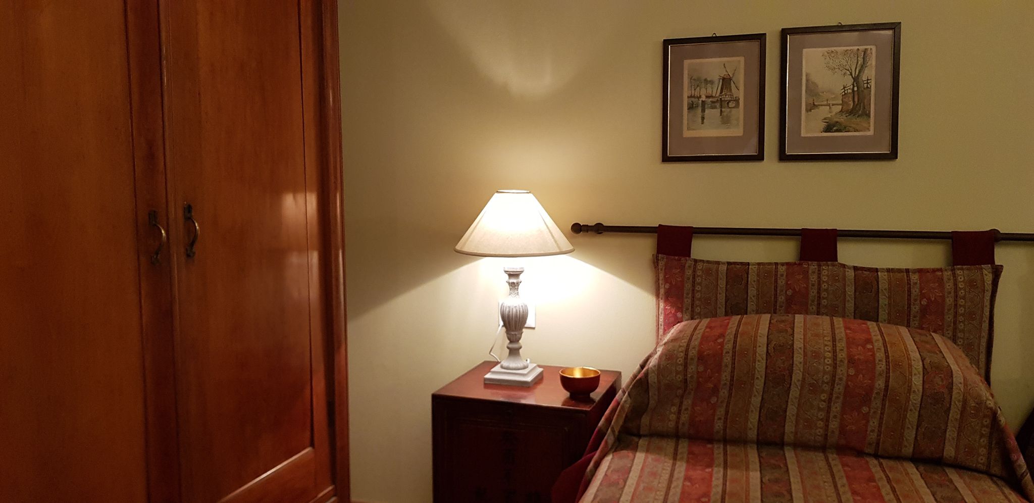 A hotel room with a wooden wardrobe, a bedside table with a lamp, a bed with striped bedding, and two framed pictures on the wall.