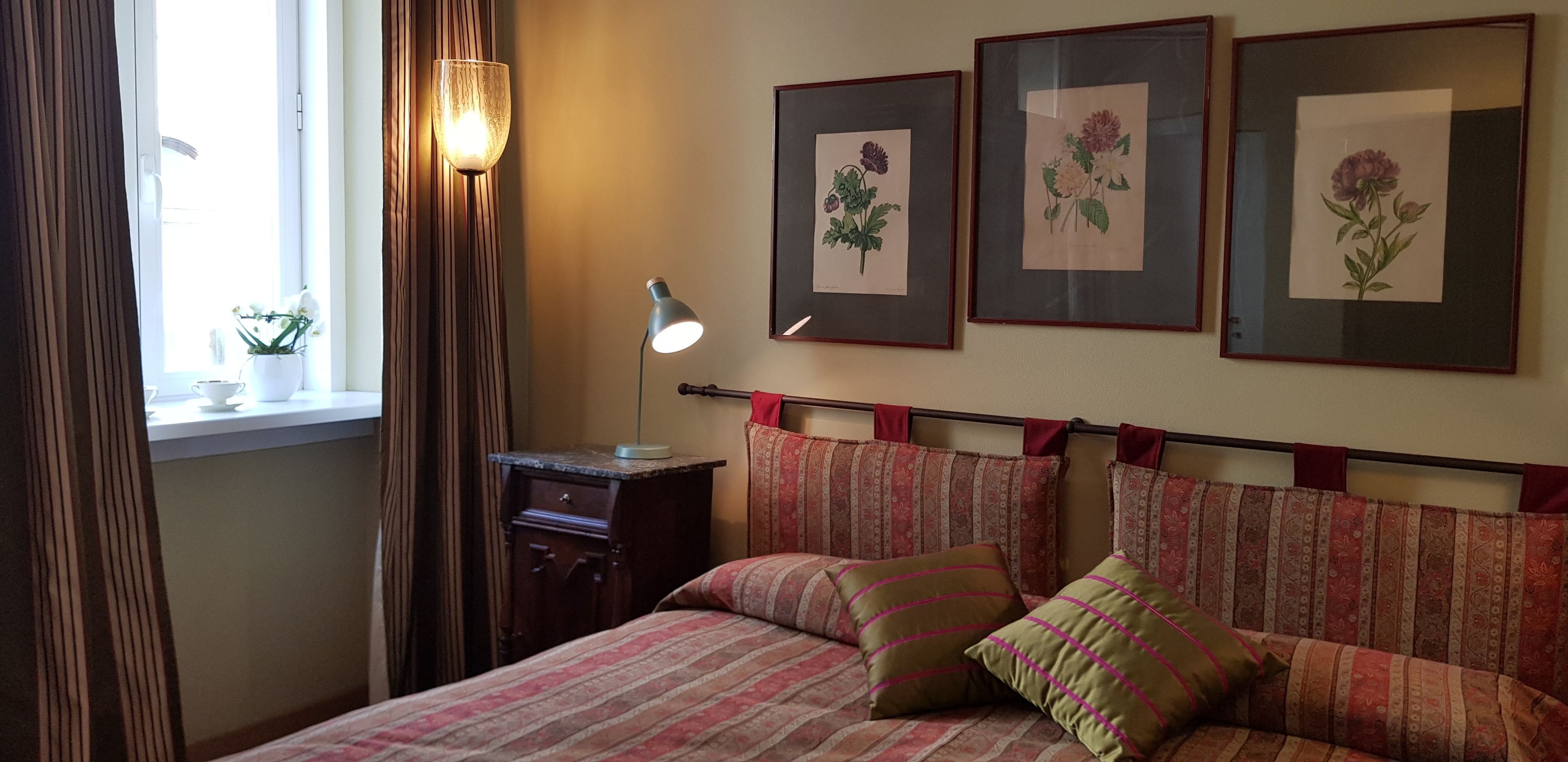 A bedroom with a bed, a nightstand, a lamp, and three framed floral artworks on the wall.