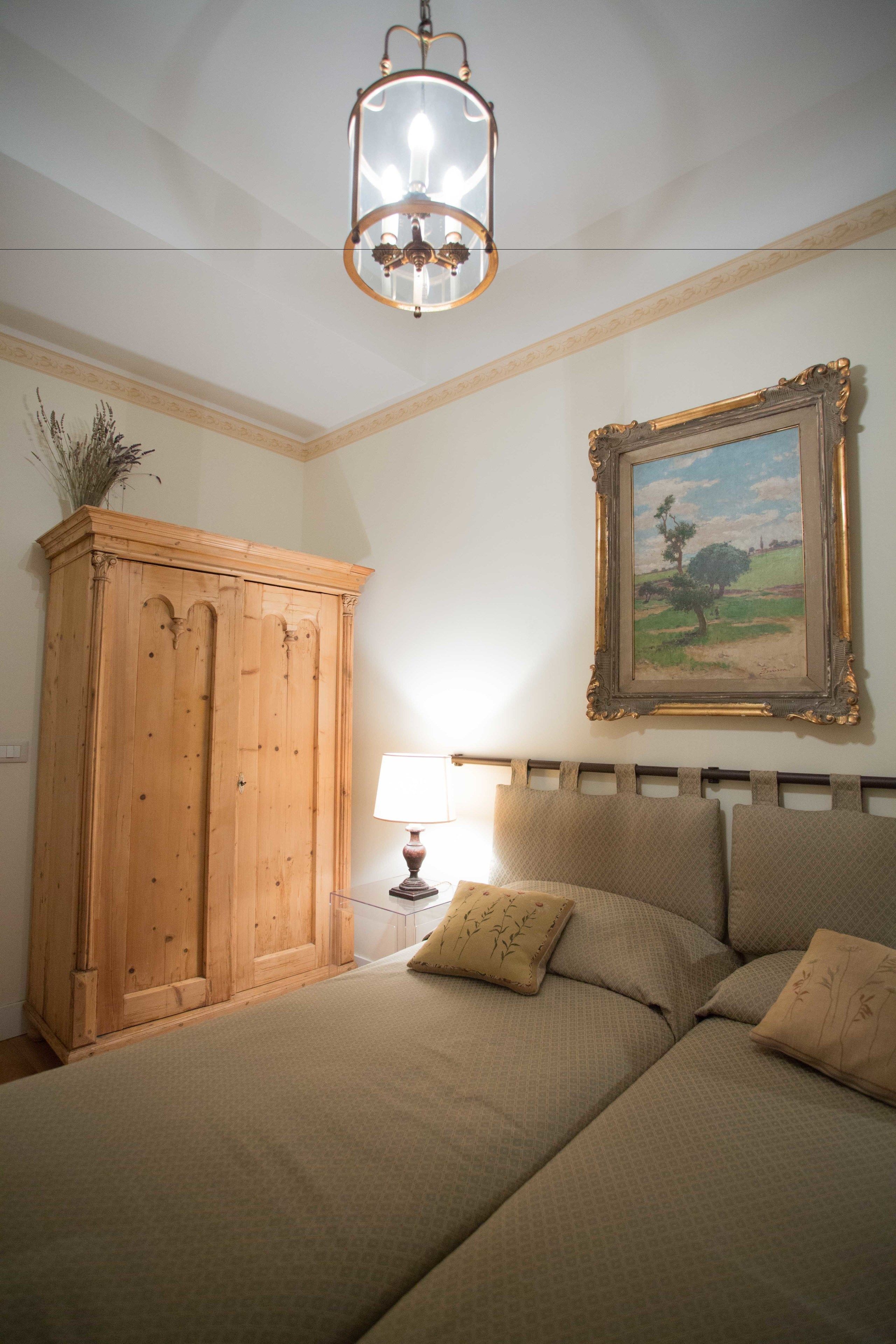 A bedroom with a bed, a wooden wardrobe, a painting, and a chandelier.