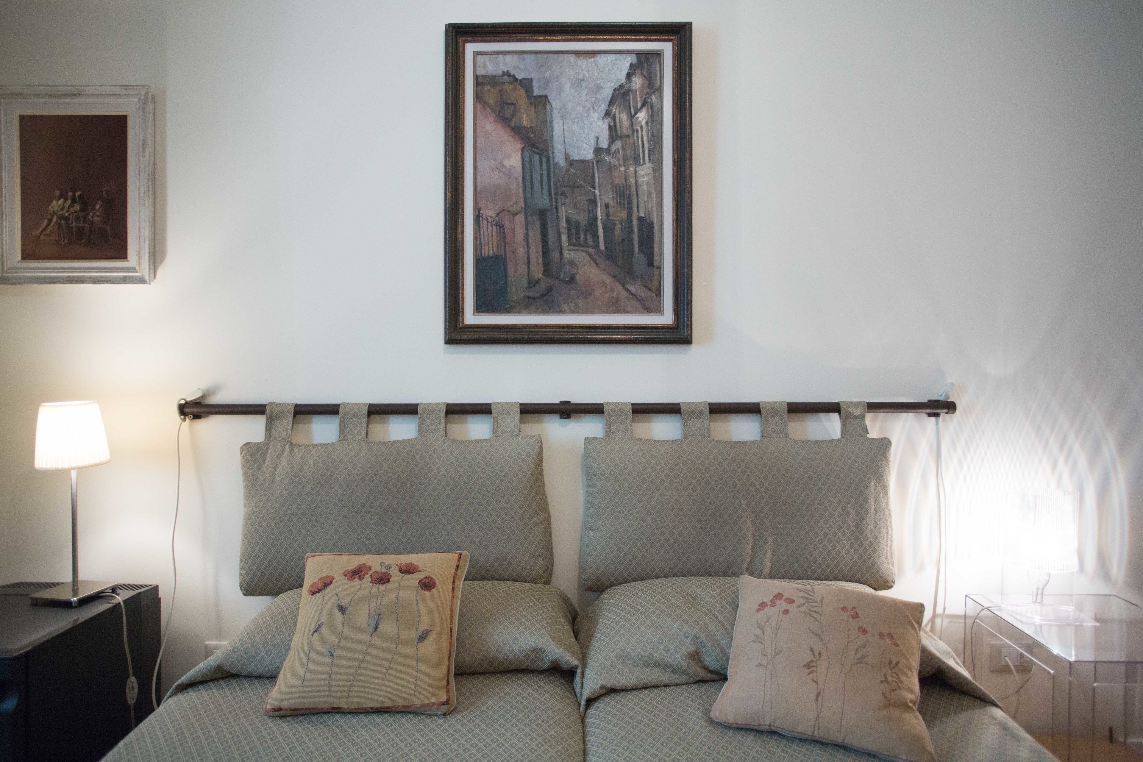 A bedroom with a bed, pillows, a nightstand, a lamp, a painting, and a window.