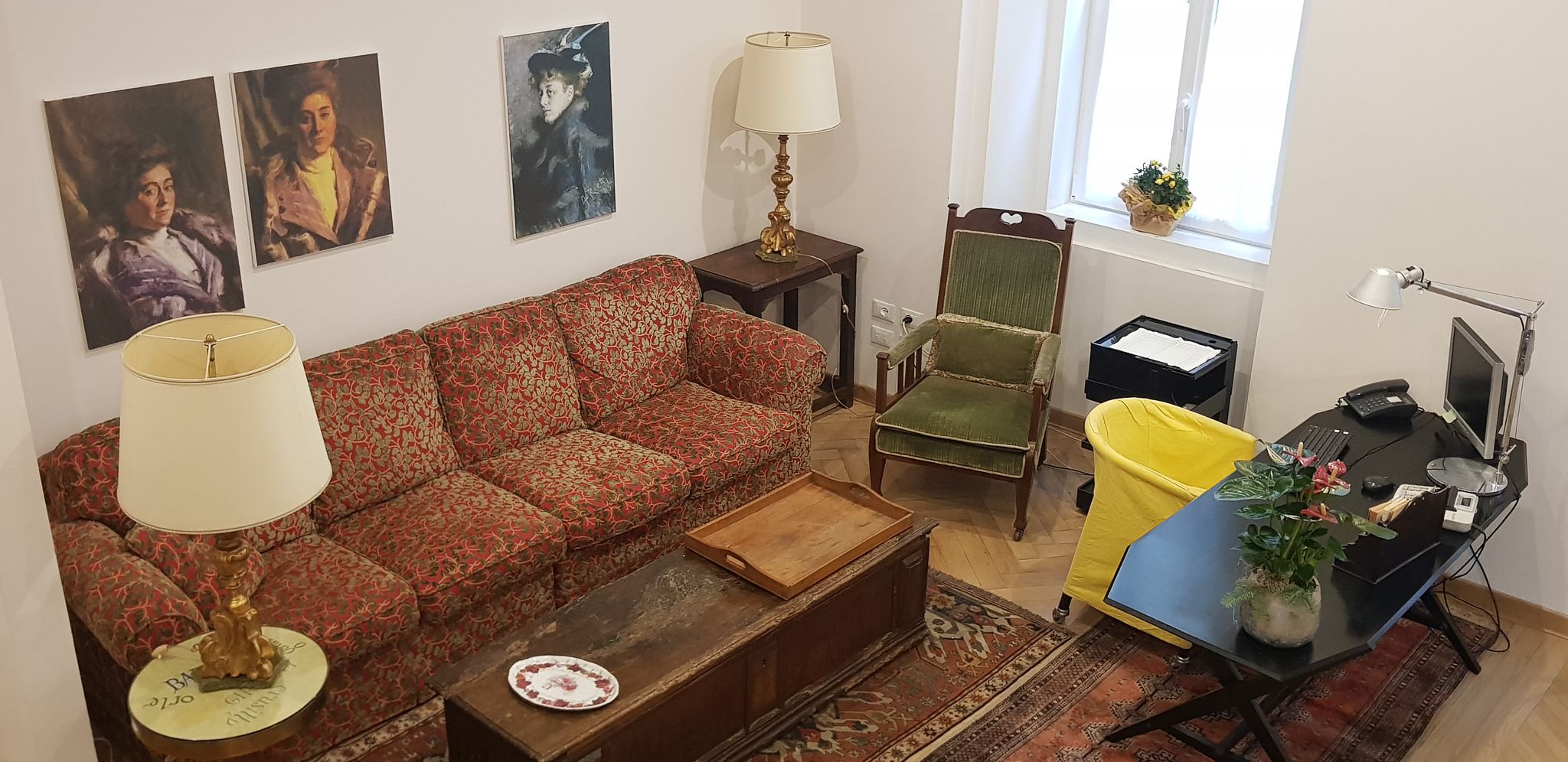 A living room with a sofa, two chairs, a coffee table, a desk with a computer, and a window with a plant.