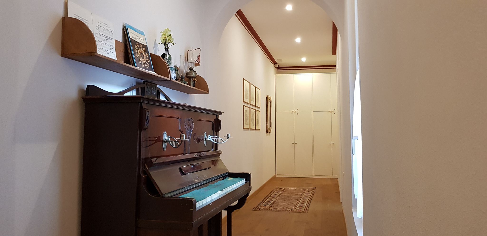 Un pasillo con un piano, una estantería, cuadros enmarcados y una puerta.