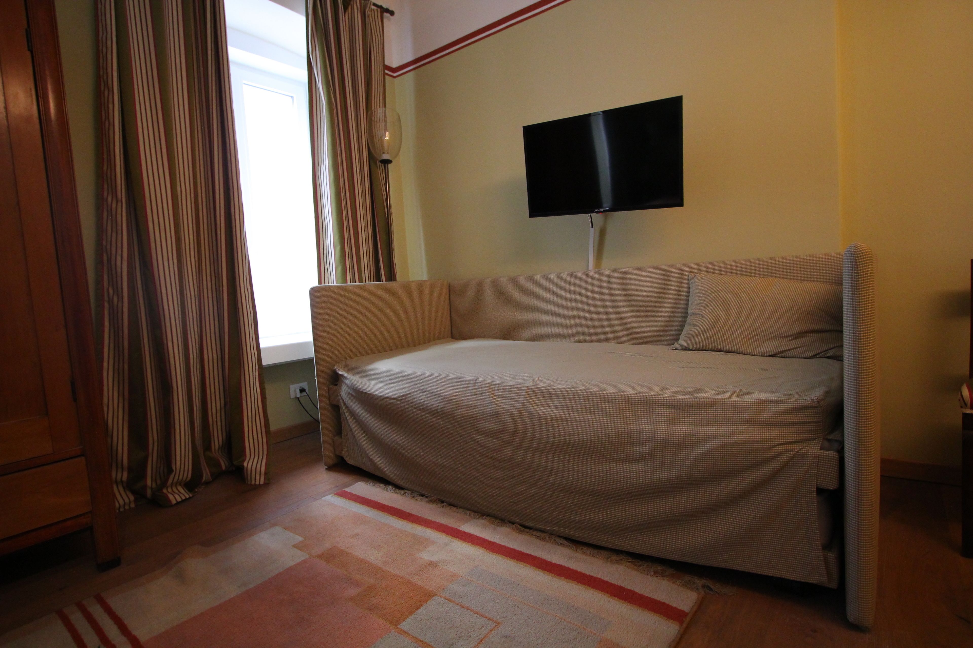 Uma cama de solteiro com protetor de colchão, uma televisão fixada na parede, uma janela com cortinas e piso de madeira.