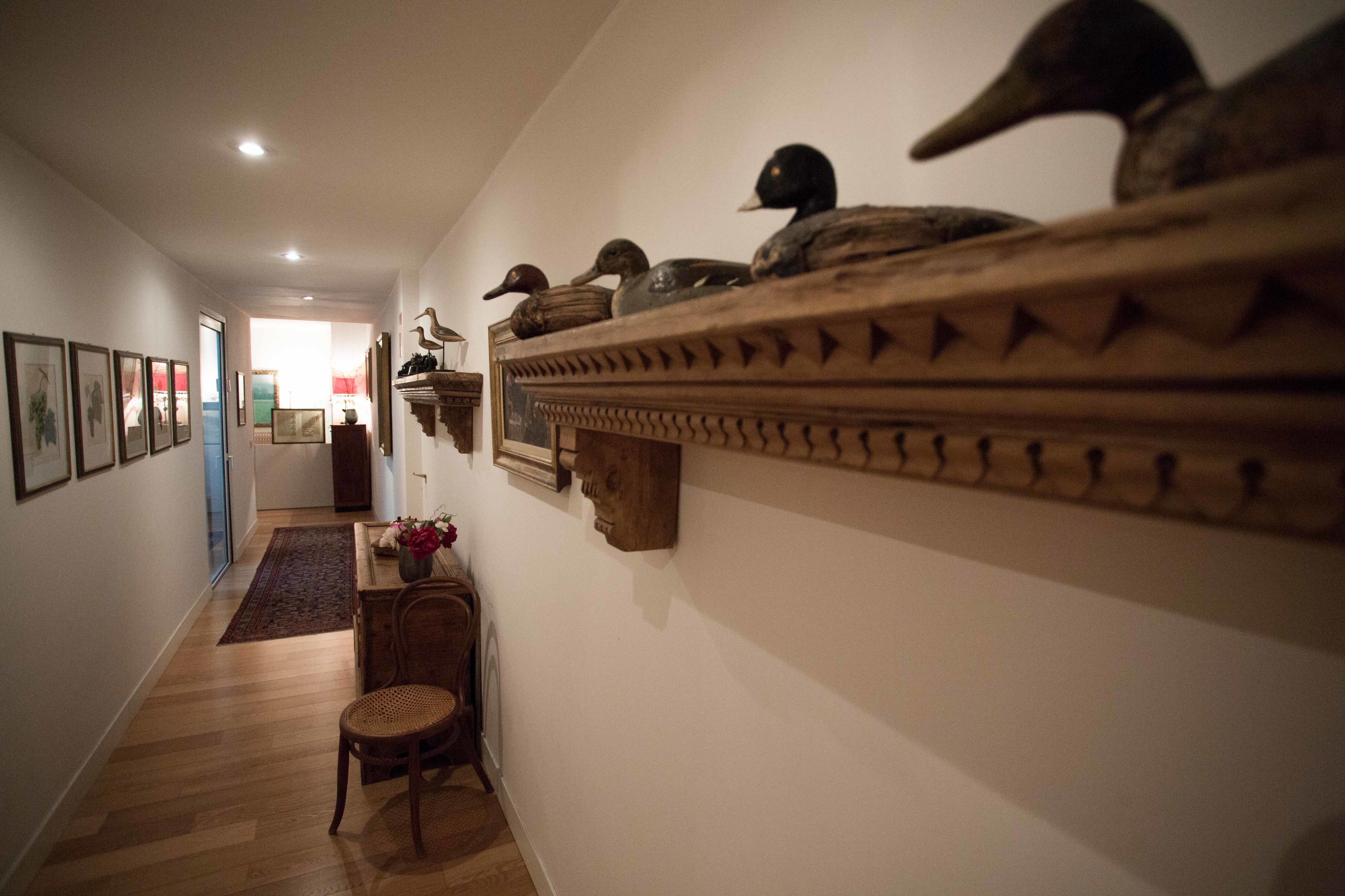 Un couloir doté d’un plancher en bois, d’une chaise, d’une étagère décorative ornée de canards et de tableaux encadrés accrochés au mur.