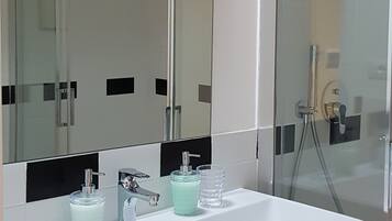 Un baño moderno con un lavamanos blanco, dos dispensadores de jabón de vidrio, un espejo grande y una mampara de ducha de vidrio.