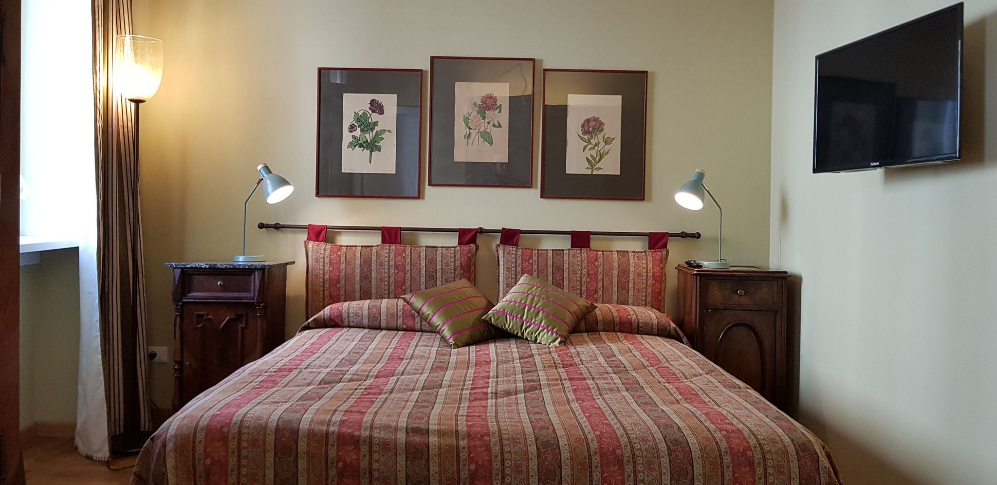 A neatly arranged bedroom with a large bed, two bedside tables with lamps, a flat-screen TV mounted on the wall, and three framed floral artworks above the bed.