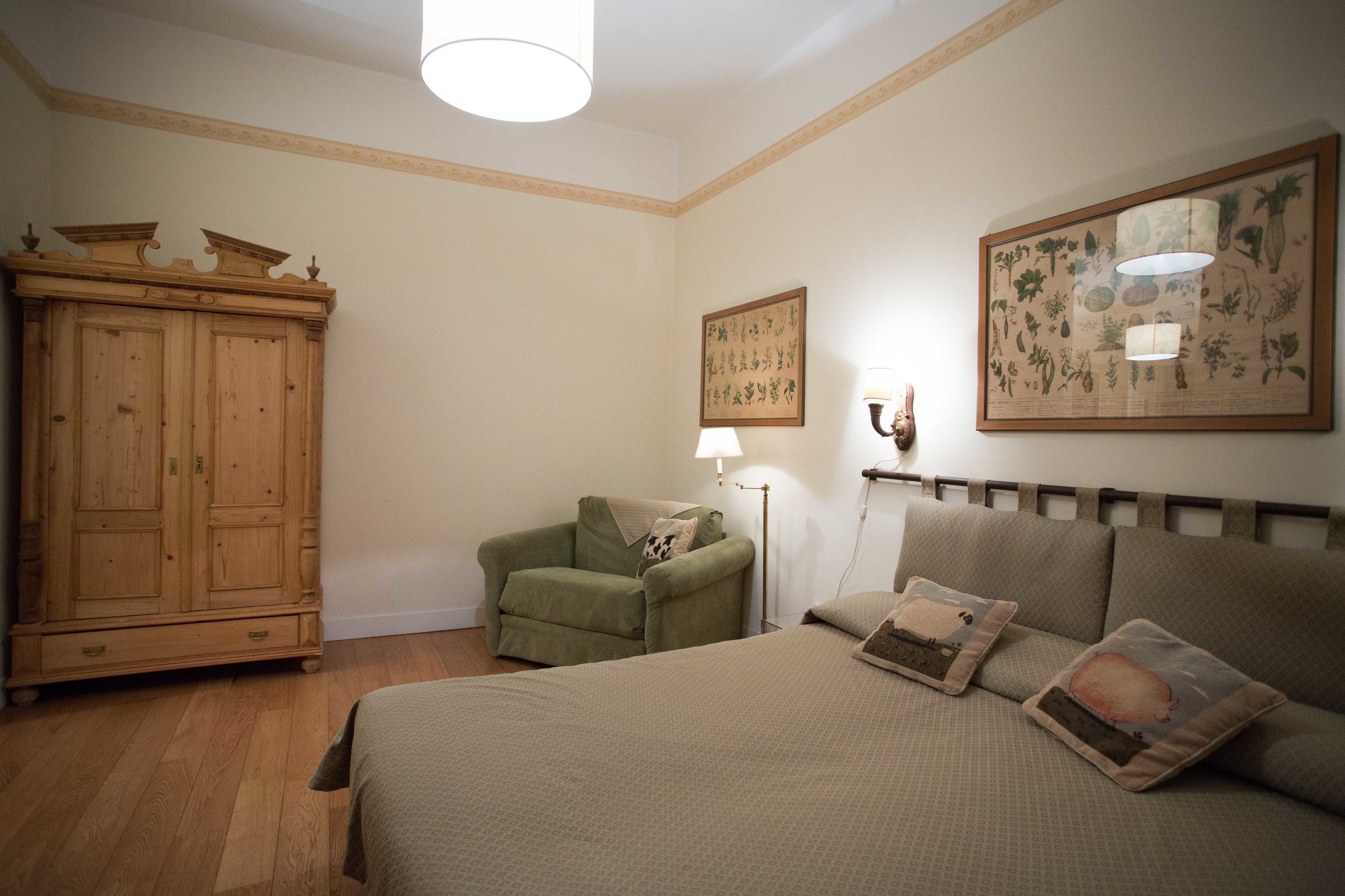 A bedroom with a bed, a green armchair, a wooden wardrobe, and two wall-mounted lamps.