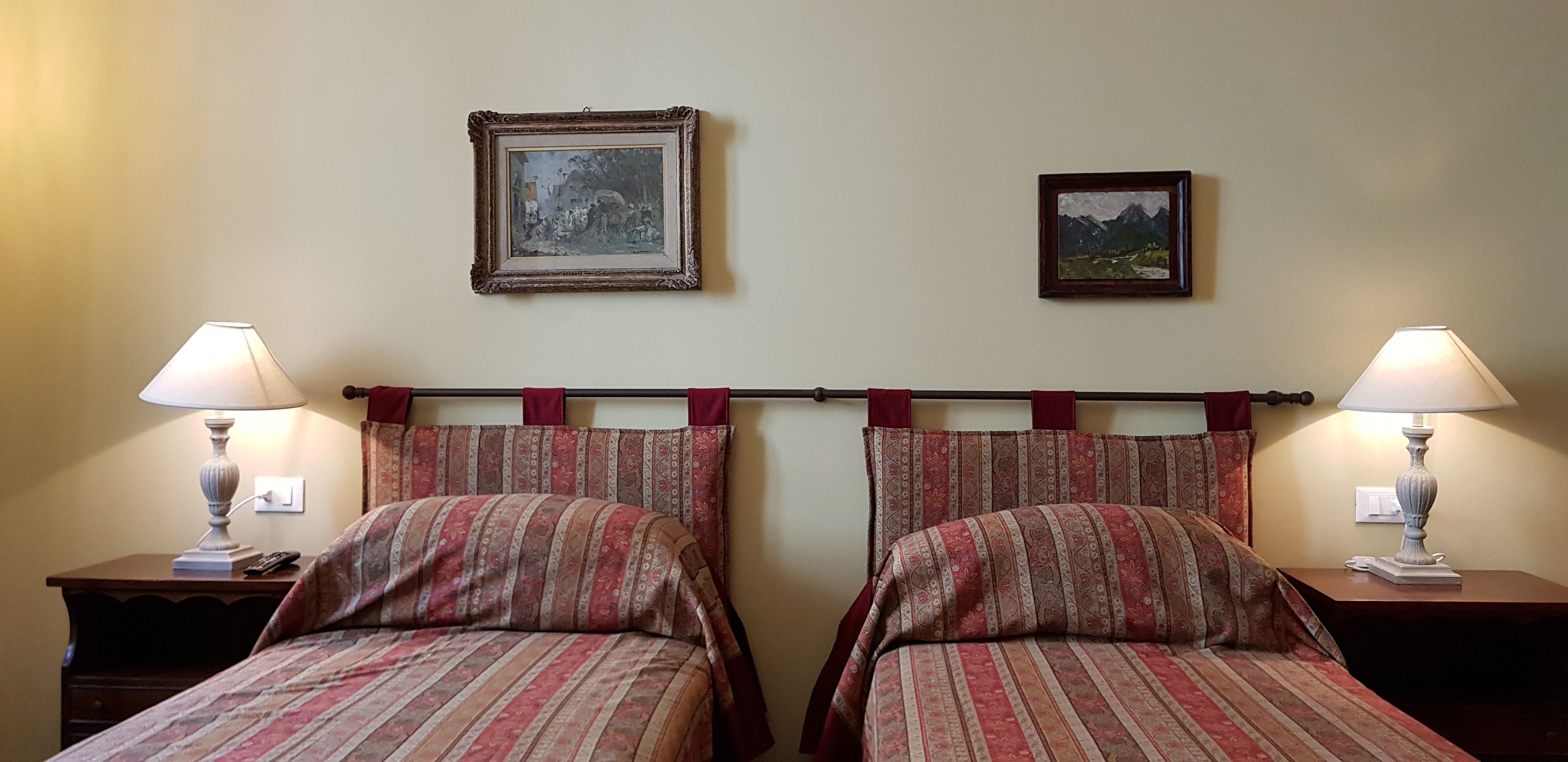Habitación de hotel con dos camas, cada una con una colcha y almohadas estampadas, flanqueadas por mesitas de noche con lámparas. Dos cuadros enmarcados decoran la pared sobre las camas.