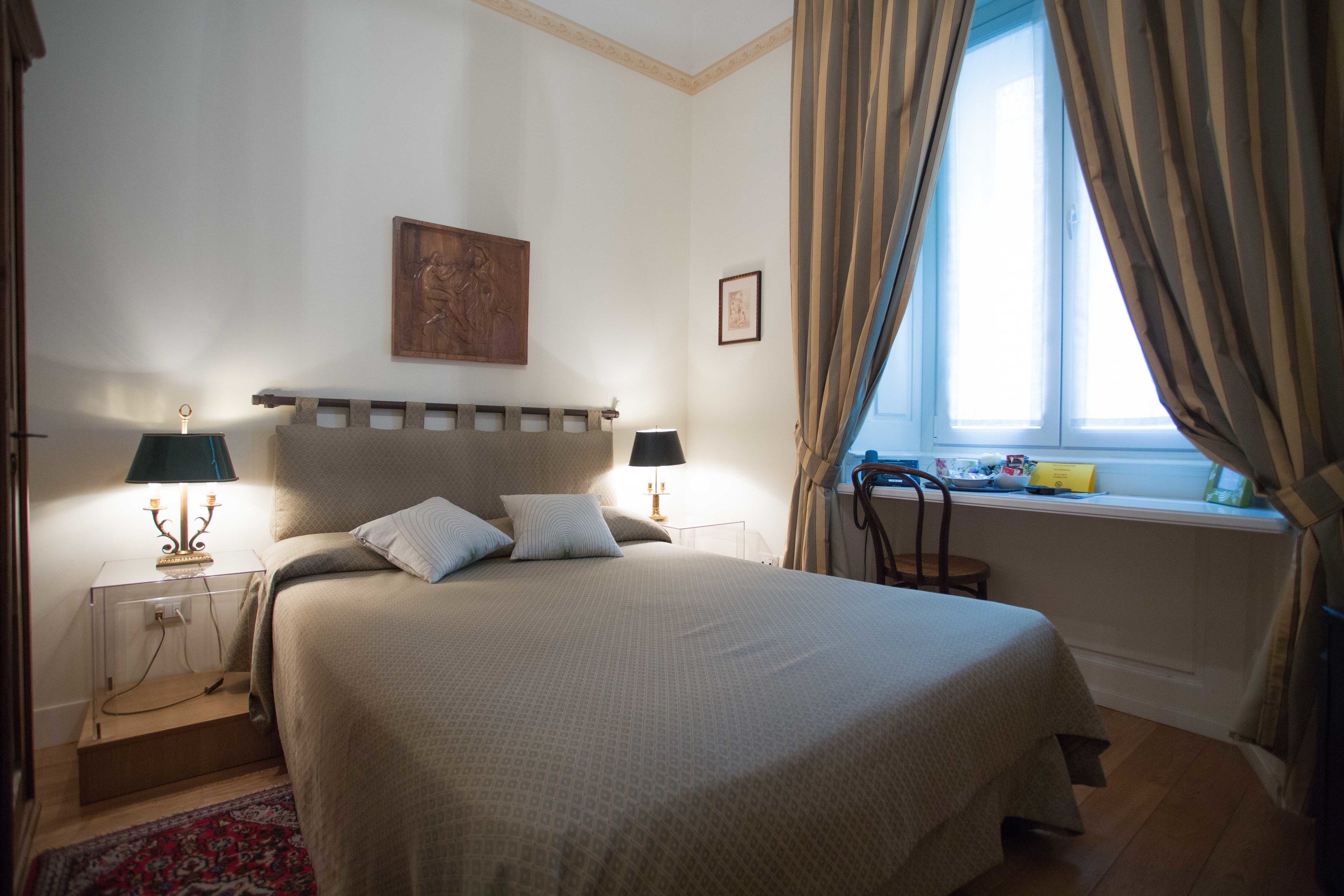 Un dormitorio ordenado con cama, lámparas de noche, ventana con cortinas y una mesita con lámpara.