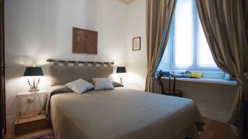 Un dormitorio ordenado con cama, lámparas de noche, ventana con cortinas y una mesita con lámpara.