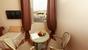 A small room with a bed, a table with fruit, a chair, and a view of buildings through a window. - Residenza Gens Julia (Rome)