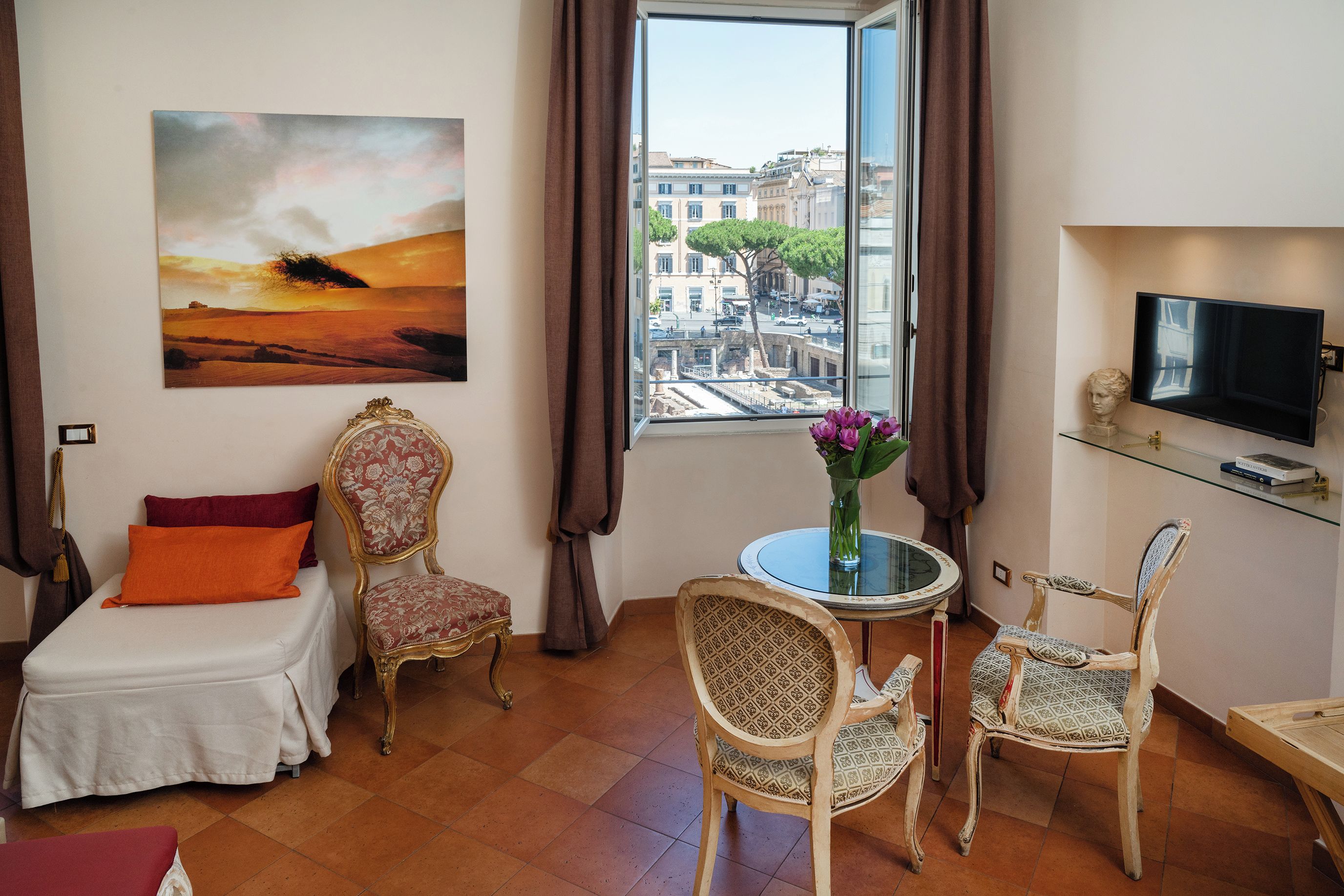 A hotel room with a bed, two chairs, a small table with a vase, a painting, and a TV on the wall.