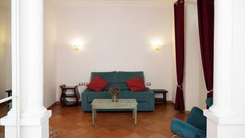 Un salon avec un canapé de couleur turquoise, deux coussins rouges, une petite table sur laquelle est posé un vase de fleurs et un fauteuil bleu.
