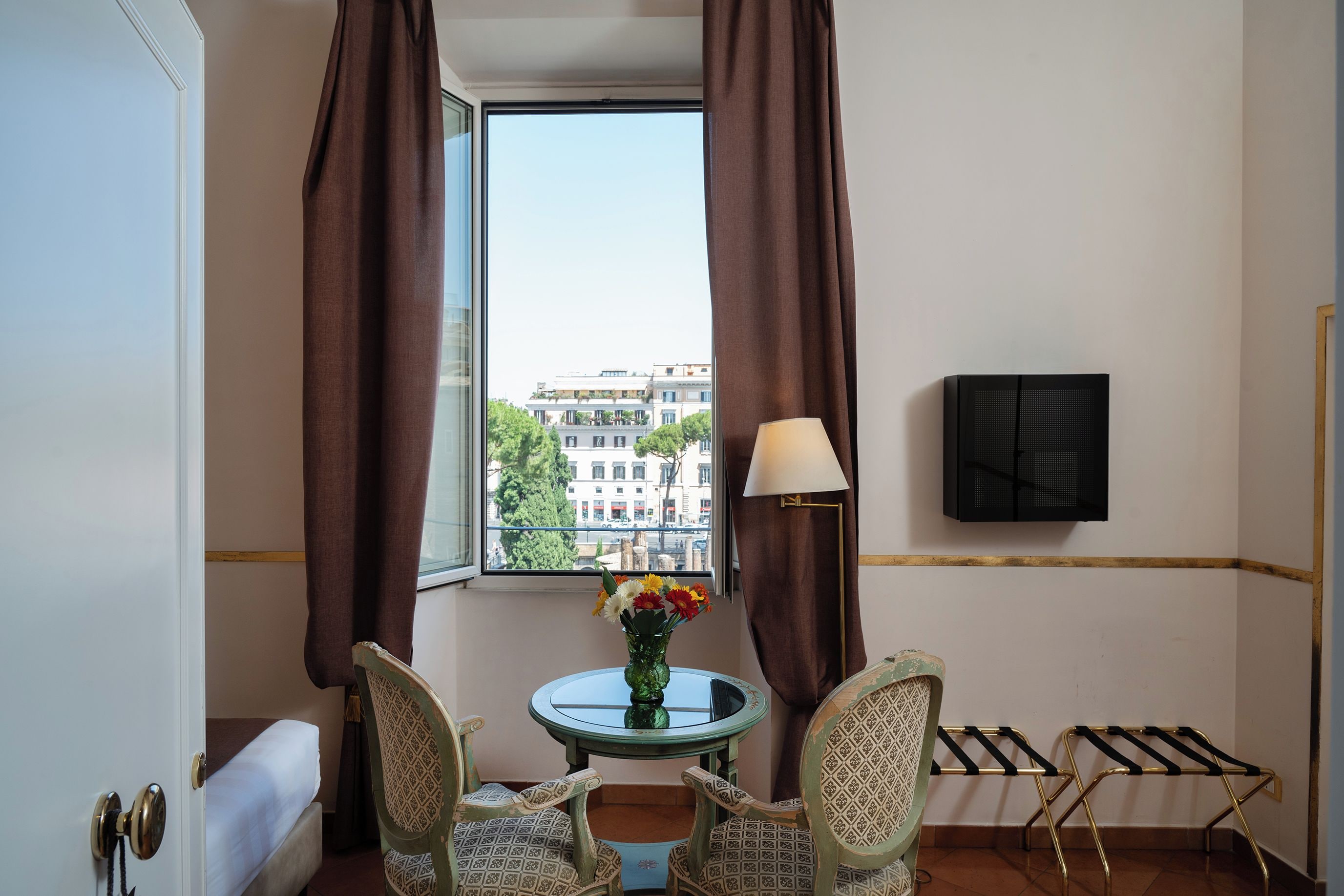 A hotel room with a bed, a small table with a vase of flowers, two chairs, a window with curtains, a lamp, and a radiator.