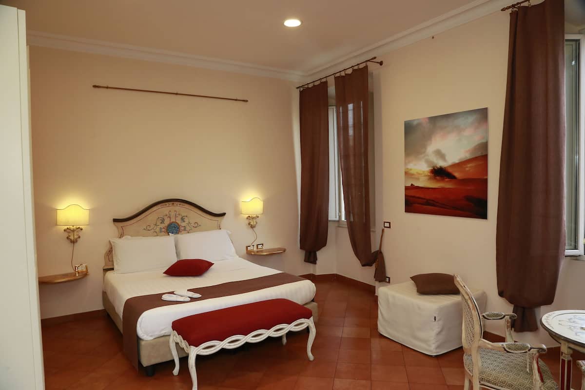 A hotel room with a bed, bedside lamps, a small table, a chair, and a painting on the wall.