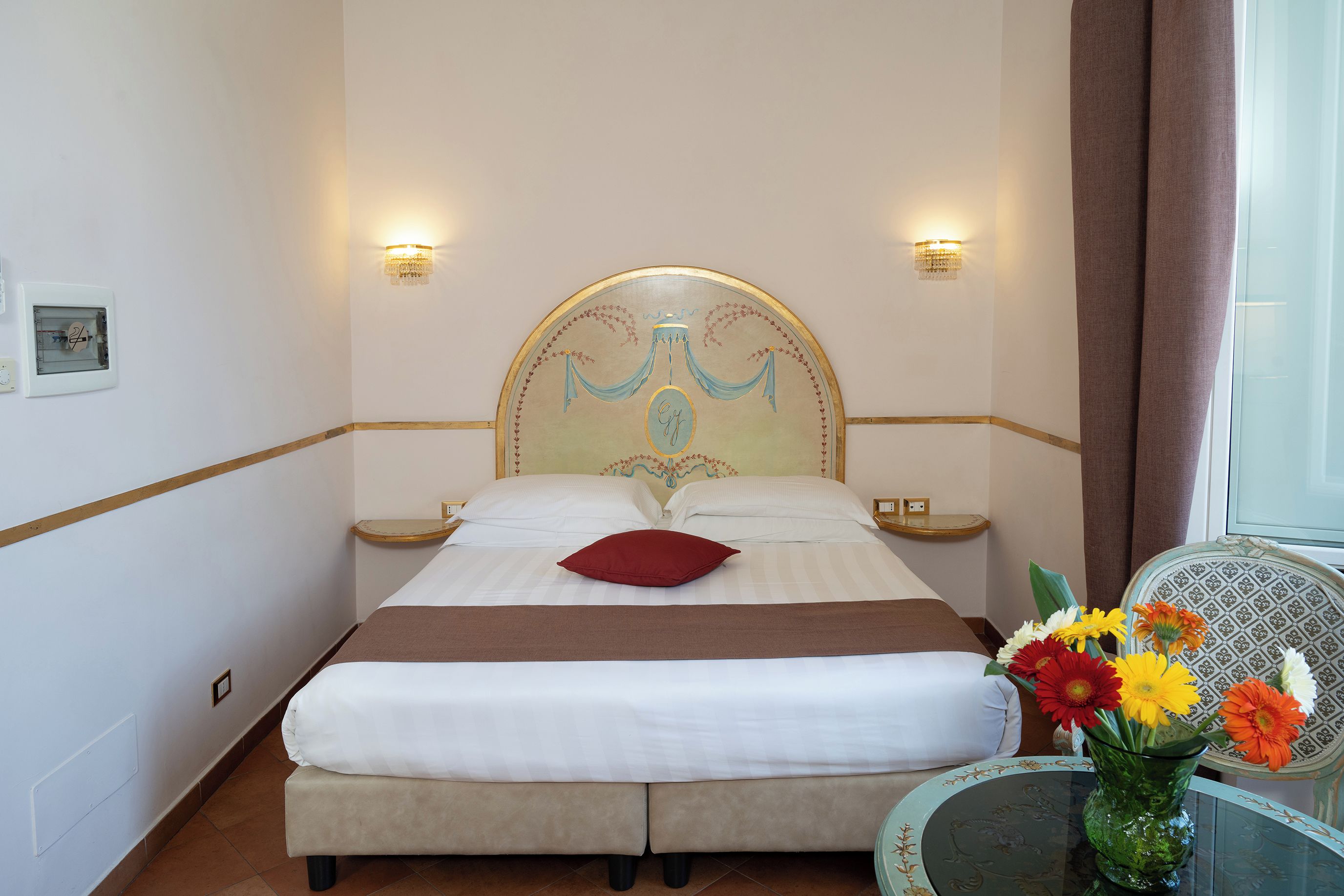 Een hotelkamer met een tweepersoonsbed, twee nachtkastjes, een klein tafeltje met een bloevas en een raam met gordijnen.