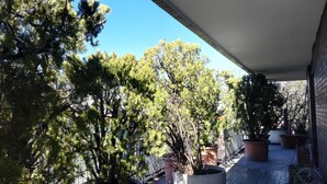 Un balcón con plantas en macetas y árboles, cielo azul despejado y una pared de ladrillo.
