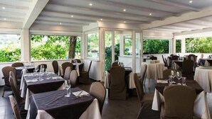 A restaurant with multiple tables and chairs, large windows with a view of greenery, and a modern ceiling design. - Villa Fernanda (Massa Lubrense)