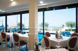 A restaurant with tables and chairs, large windows, and a view of the street and beach. - La Fiorita (Rimini)