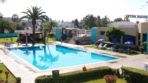 A swimming pool with lounge chairs and umbrellas, surrounded by greenery and a building with a sign. - Mabrouk Hotel and Suites- Adult Only (Agadir)
