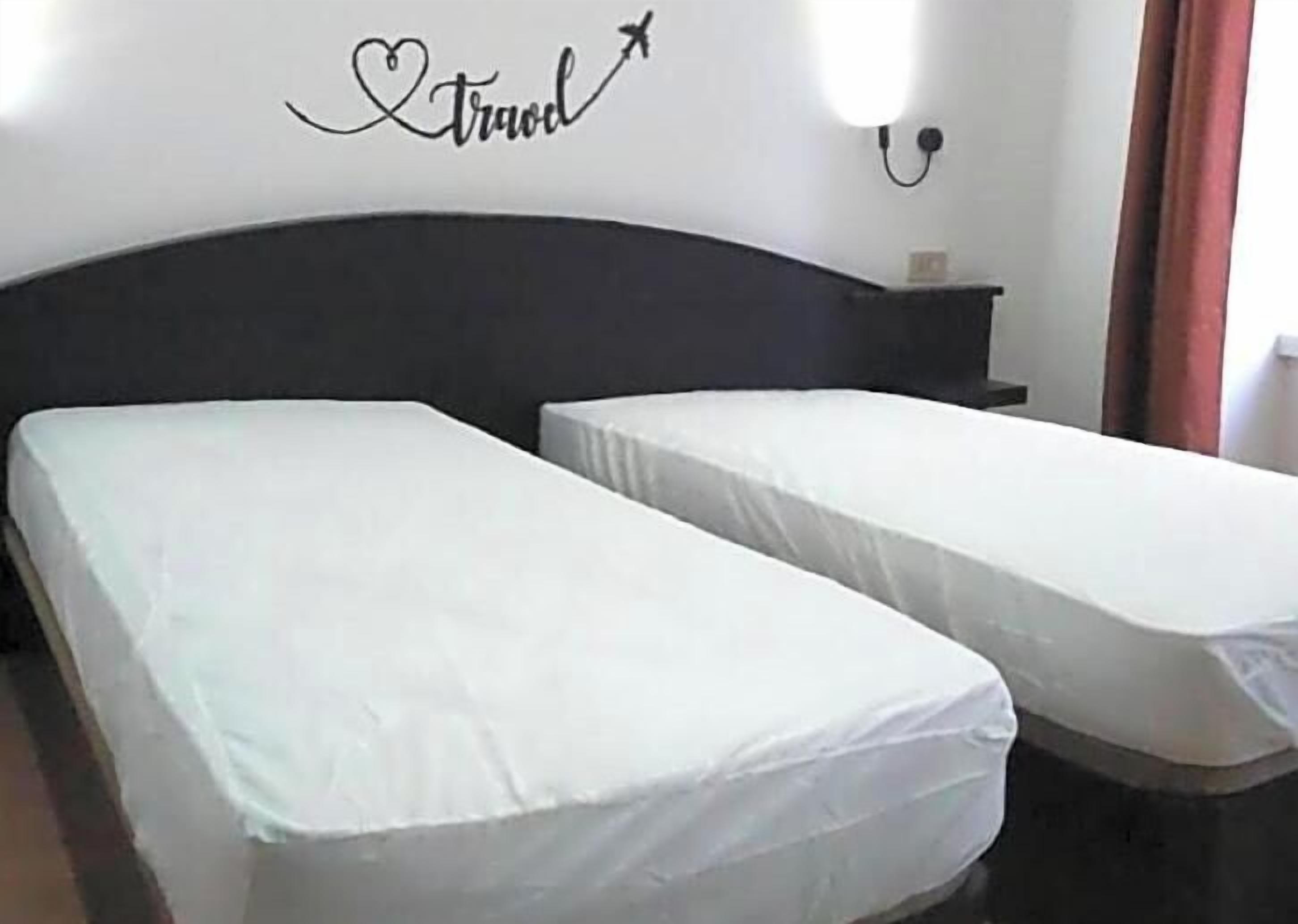 Una habitación con dos camas individuales, un cabecero oscuro, una lámpara fijada a la pared y un adorno con la palabra 'travel' y un corazón.