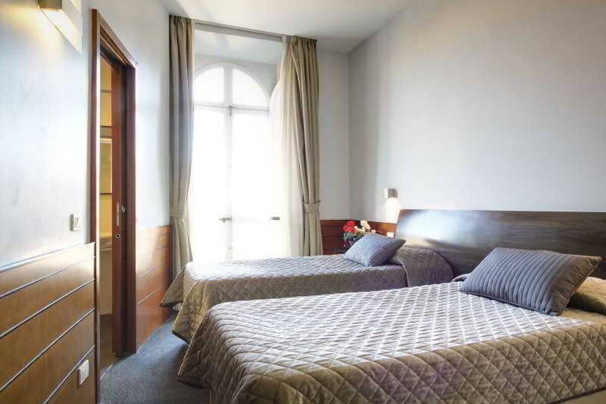 Hotellihuone, jossa on kaksi sänkyä, puinen vaatekaappi, ikkuna, jossa on verhot, ja yöpöytä, jossa on lamppu.